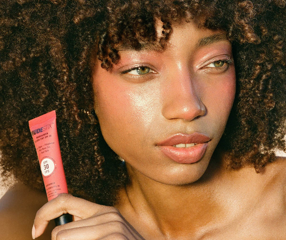 Dark skinned young woman holding a tube of Nudescreen Blush