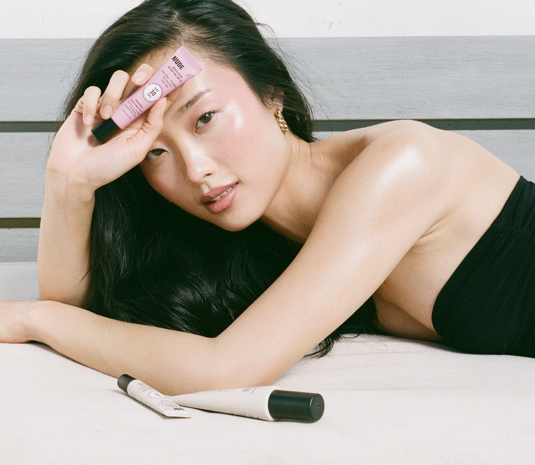 Young Asian woman laying on her side while holding a tube of Nudescreen blush tint